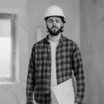 A construction worker with a hard hat and plaid shirt holding a blueprint inside a renovation site.