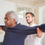 A senior man is assisted by a caregiver for stretching exercises indoors.